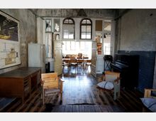 Ein rustikaler Innenraum mit Holzboden, freiliegenden, teils verputzten Backsteinwänden, und einer Einrichtung aus hölzernen Stühlen, Tischen und einem schwarzen Klavier. Im hinteren Bereich des Raums befinden sich große Fenster und doppeltürige Durchgänge, die ein sonnenbeschienenes Esszimmer sichtbar machen.