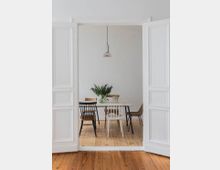 Ein minimalistischer Essbereich mit einem hellen Holzboden, einem weißen Esstisch, verschiedenen Holzstühlen und einer Vase mit Blumen. Die Szene wird von einer Pendelleuchte und weißen, geöffneten Doppeltüren eingerahmt.