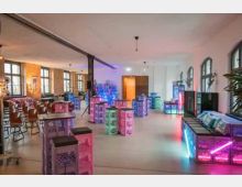Großer, heller Innenraum mit hohen Decken, Stützsäulen und Bogenfenstern, eingerichtet mit Tischen, Stühlen und modularen Sitz- und Ablageelementen aus Kisten. Farbiges Neon- und LED-Licht beleuchtet einzelne Bereiche, mit Bar- bzw. Loungebereich entlang der Wand.