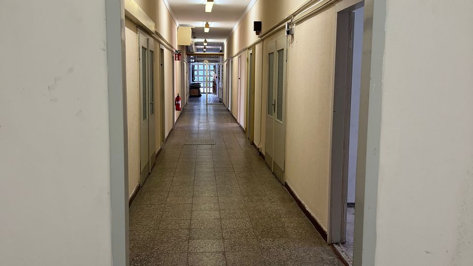 Langer, schmaler Innenflur eines Gebäudes mit Fliesenboden, hellen Wänden und mehreren Türen auf beiden Seiten, beleuchtet durch Deckenleuchten. Am Eingang hängt ein grünes Notausgangsschild, am Ende des Gangs ist eine verglaste Tür zu sehen.