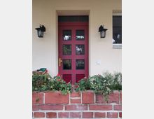 Rote Eingangstür mit gläsernen Fenstern in dunklen Rahmen, flankiert von zwei schwarzen Wandlaternen. Davor steht ein niedriger Ziegelstein-Pflanzkasten mit grünem Blattwerk und roten Blüten.