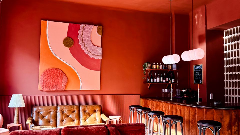 Warm-toned lounge bar interior with red walls, a coffered ceiling and pendant lights, featuring plush velvet seating and a round coffee table in the foreground. A wooden bar counter with tall stools, shelves of bottles, and a wall-mounted chalkboard menu sits along the right side.