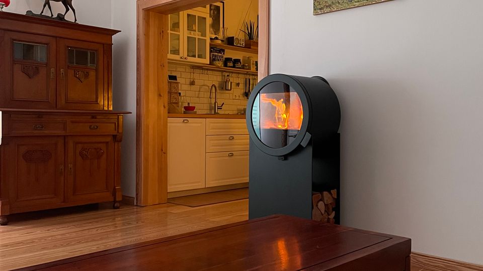 Wohnzimmer mit Holzboden, großem dunklen Couchtisch und einem modernen schwarzen Kaminofen mit Sichtfenster; daneben steht ein antiker Holzschrank. Durch einen breiten Türrahmen ist eine helle Küche mit weißen Schränken und Fliesenspiegel zu sehen, an der Wand hängt ein Gemälde.