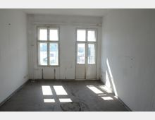 Leerer, lichtdurchfluteter Raum mit weißen Wänden, zwei großen Fenstern und einer Glastür, die zu einem Balkon führt. Der Boden ist aus Beton, und an den unteren Wänden sind alte Rohrleitungen sichtbar.