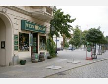 Außenansicht eines Weinladens namens "Wein Oase" mit grüner Beschilderung und großen Fensterfronten, die viele Flaschen ausstellen. Vor dem Laden stehen Holzfässer, Pflanzen und ein Werbeschild, umgeben von einem gepflasterten Gehweg und einer Straßenkreuzung mit Bäumen im Hintergrund.