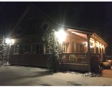 Ein eingeschossiges Holzhaus mit Veranda, das bei Nacht beleuchtet ist. Der Vorderbereich und das Dach sind teilweise schneebedeckt, und warme Lichter schmücken die Hausfront.