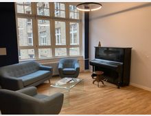 Ein kleiner, gemütlicher Raum mit Holzboden, einer Sitzgruppe aus zwei Sesseln, einem Sofa in blauem Stoff und einem gläsernen Couchtisch. Im Hintergrund steht ein schwarzes Klavier mit einem Hocker, vor großen Fenstern, die den Blick auf die Fassade eines Backsteingebäudes bieten.