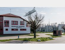 Ein Gebäude der Freiwilligen Feuerwehr Treuenbrietzen mit weißer Fassade, roten Fensterrahmen und einem rot gedeckten Dach steht links im Bild. Rechts davon befindet sich ein Umspannwerk mit zahlreichen Stromleitungen und technischen Anlagen, umgeben von einer Grünfläche mit vereinzelten Bäumen.