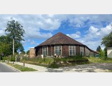 Holzgebäude mit achteckigem Grundriss, großen Sprossenfenstern und einem roten Ziegeldach, umgeben von einem Zaun und überwucherter Vegetation. Im Vordergrund verlaufen ein Gehweg und eine Straße, während Bäume und blauer Himmel die Umgebung prägen.