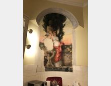 Wandbild in einer Nische mit gemaltem Rundbogen, das eine antik anmutende Szene zeigt: Eine stehende Frau in einem langen Kleid und ein Engel in einer Gartenszenerie mit Skulpturen und Zypressen im Hintergrund. Darunter befinden sich moderne Küchengeräte und weiße Wandfliesen.