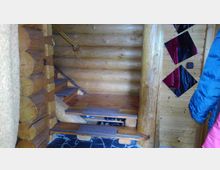 Holzinterieur mit einer kleinen Treppe aus massiven Holzbrettern, einem Geländer an der Wand und einer Verkleidung aus Rundholz. Rechts ist eine Wand mit diagonalen Spiegeln und eine Person im blauen Mantel teilweise sichtbar.