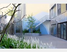 Moderner Innenhof mit bepflanzten Beeten, einer Wand mit blauer geometrischer Bemalung und einer schmalen Metalltreppe. Die umliegenden Gebäude haben Fassaden mit verglasten Fenstern und glänzenden Metallflächen.
