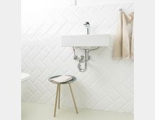 Ein modernes Badezimmer mit weißen, diagonal angeordneten Fliesen an der Wand. Ein kleines weißes Waschbecken mit sichtbaren verchromten Rohren, daneben beige Handtücher an einem Haken und ein kleiner, goldener Beistelltisch mit einem Handtuch darauf.