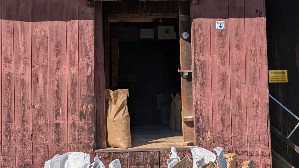 Alte Scheune mit verwitterter Holzfassade und offener Tür, die in einen dunklen Innenraum führt. Davor steht auf einer Rasenfläche ein kleiner Anhänger mit mehreren weißen und braunen Säcken.