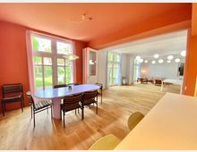 Ein heller Raum mit Parkettboden, unterteilt in zwei Bereiche: Ein Essbereich mit einem ovalen Tisch und Stühlen vor hohen Fenstern mit Gartenblick, und ein angrenzender Wohnbereich mit modernen Sitzmöbeln und kugelförmigen Hängeleuchten. Wände sind teils in Rot und teils in Weiß gehalten.