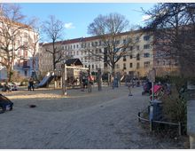 Ein städtischer Kinderspielplatz mit Sandboden, Klettergerüsten, Schaukeln und einer Rutsche, umgeben von mehrstöckigen Altbauwohnhäusern. Bäume ohne Laub und parkende Kinderwagen sind im Vordergrund zu sehen, während Kinder und Erwachsene den Spielplatz nutzen.