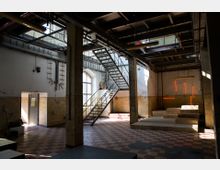 Industriehalle mit hohen Decken, einer Metalltreppe, großen Fensterflächen und sichtbaren Rohrleitungen. Der Boden ist mit rot-beigen Fliesen gestaltet, und an einer Wand befinden sich leuchtende orangefarbene Lichtinstallationen.