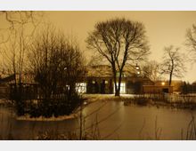 Nachtszene eines verschneiten Gartens mit gefrorenem Teich im Vordergrund, Bäumen und Sträuchern, sowie einem großen Gebäude mit erleuchteten Fenstern im Hintergrund. Der Himmel ist durch künstliches Licht rötlich-orange gefärbt.