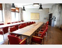 Industrieller Besprechungsraum mit langen Holztischen, umgeben von roten Polsterstühlen. Die Wände sind unverputzt, große Fenster lassen Tageslicht herein, und an der Wand hängt eine Karte neben alten Schaltkästen.