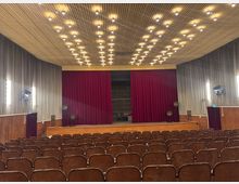 Ein Theater- oder Veranstaltungssaal mit einer Bühne, vor der ein schwerer, roter Vorhang hängt. Der Saal hat viele braune Sitzreihen, eine holzvertäfelte Wandverkleidung und eine Decke mit symmetrisch angeordneten, warm leuchtenden Deckenleuchten.