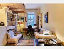 Ein gemütliches Zimmer mit Hochbett aus Holz, einer Leiter, einem Schreibtisch vor einem großen Fenster mit weißen Vorhängen sowie einem Sofa mit Kissen. Regale, Bücher und persönliche Dekorationsgegenstände füllen den Raum, der von warmem Licht beleuchtet wird.