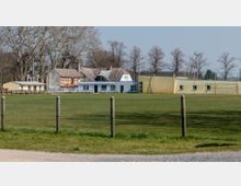 Eine Grünfläche mit einem Sportplatz, umgeben von einem Drahtzaun, im Hintergrund stehen Gebäude mit Ziegeldächern und Fachwerk, daneben eine gelbliche Wand mit kleinen Fenstern und ein Tor. Im Hintergrund befinden sich kahle Bäume und ein klarer Himmel.