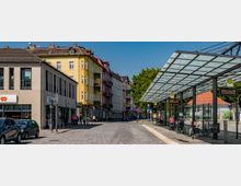 Städtische Straßenszene mit einer modernen Bushaltestelle mit Glasüberdachung und Sitzbereichen auf der rechten Seite. Links befinden sich mehrstöckige Wohn- und Geschäftsgebäude, darunter ein gelbes Gebäude und Geschäfte wie eine Bäckerei und eine Drogerie; geparkte Autos sowie einige Passanten prägen das Bild.