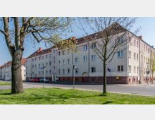 Mehrstöckiges Wohngebäude im Stil der Nachkriegsarchitektur mit beige verputzten Fassaden, rotbraunem Sockelbereich und roten Satteldächern, flankiert von Bäumen und einer angrenzenden Straße mit geparkten Autos. Im Vordergrund eine Grünfläche mit zwei schlanken Bäumen.