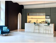 Moderne offene Küche mit dunklen Schränken, einer beleuchteten Rückwand und einer zentralen Kücheninsel aus hellen Steinplatten. Im Vordergrund stehen zwei Barhocker, daneben ein blauer Sessel, und links ist eine weiße Wand mit einem großen Fenster sichtbar.