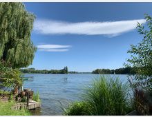 Ein See mit klarem Wasser, umgeben von dichtem Grün und Bäumen, darunter eine große Trauerweide am linken Bildrand. Im Vordergrund befinden sich Uferpflanzen, Blumentöpfe und eine kleine gemütliche Terrasse, während der Himmel blau ist mit einzelnen Wolken.