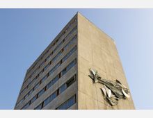 Hohes, rechteckiges Betongebäude mit mehreren Stockwerken und schmalen Fenstern, an dessen Außenwand eine abstrakte Metallskulptur angebracht ist; blauer Himmel im Hintergrund.