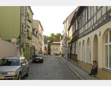 Pflastersteinstraße mit eng stehenden Gebäuden auf beiden Seiten, darunter Fachwerk- und Putzfassaden; im Hintergrund ist ein großes Fachwerkhaus zu sehen. Links ein grünes Gebäude mit Hotel-Schriftzug, rechts Menschen vor einem Eingang sitzend.