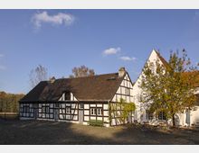 Fachwerkhausensemble mit weiß verputzten Wänden und schwarzen Holzbalken in ländlicher Umgebung, gepflasterter Hof mit Tischen und Stühlen sowie einem Baum im Herbstlaub. Im Hintergrund sind Bäume und ein klarer Himmel zu sehen.