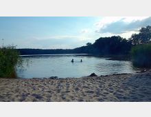 Sandstrand mit Blick auf einen ruhigen See, in dem zwei Personen im Wasser stehen; dichte Ufervegetation mit Schilf und Wald erstreckt sich im Hintergrund.