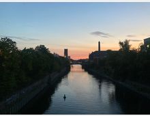 Kanal bei Sonnenuntergang, umgeben von Bäumen auf der linken Seite und industriellen Backsteingebäuden mit einem Schornstein auf der rechten Seite. Im Hintergrund ist eine Brücke und ein Turm zu sehen, der Himmel zeigt Farbtöne von Rosa, Orange und Blau.