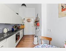 Schmale Küche mit weißen Einbauschränken und grauer Arbeitsplatte, Herd und Spüle auf der linken Seite. Holzdielenboden, eine Tür mit Glaseinsatz im Hintergrund, sowie ein Wandregal und ein runder Esstisch mit gemusterter Tischdecke auf der rechten Seite.