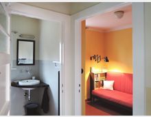 Blick durch einen Türrahmen auf zwei Räume: links ein kleines Badezimmer mit Waschbecken, Spiegel und weißen Wänden, rechts ein Wohnzimmer mit orange gestrichenen Wänden, einem roten Sofa, einer Bücherablage und einer schwarzen Wandlampe.