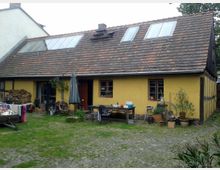 Gelbes Fachwerkhaus mit schrägem Ziegeldach sowie Dachfenstern, umgeben von einem Innenhof mit unebenem Pflaster und Rasen. Vor dem Haus stehen ein Holztisch, ein Sonnenschirm, Pflanzen in Töpfen und verschiedene Gegenstände wie Fahrräder und gestapelte Holzscheite.