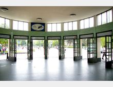 Eingangsbereich einer U-Bahn-Station mit geschwungener Fassade, grünen Wandfliesen und mehreren Glastüren, die nach außen auf einen belebten Platz führen. Oben sind große Fenster und Werbeschilder sichtbar, während draußen Menschen, Bäume und Straßenschilder zu sehen sind.