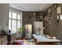 Helle Küche mit hohen Decken und einer großen Fensterfront, ausgestattet mit einer modernen Einbauküche, einem Edelstahlkühlschrank und einem langen Esstisch. An der braunen Wand hängen zahlreiche Bilderrahmen; Zimmerpflanzen und Blumen schmücken den Raum, der einen Holzboden und einen Orientteppich aufweist.