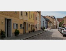 Eine ruhige, gepflasterte Straße mit mehrstöckigen Wohnhäusern in pastellfarbenen Fassaden auf der linken Seite. Rechts sind parkende Autos entlang des Gehwegs zu sehen, und Straßenlaternen säumen den Bürgersteig.