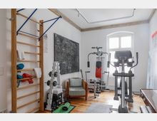 Heller Fitnessraum mit Holzboden, ausgestattet mit einer Sprossenwand, Hanteln, einer Multifunktions-Kraftstation und einem Crosstrainer. Im Hintergrund steht ein Fenster mit Rundbogenrahmen, daneben befindet sich ein Sessel und eine Staffelei.