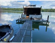 Hausboot auf einem ruhigen See, angedockt an einem schmalen, metallischen Steg. Im Vordergrund ein kleines Schlauchboot, im Hintergrund dichter Wald und ein bewölkter Himmel.
