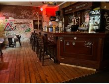 Innenraum einer urigen Bar mit langem dunklem Holztresen, Barhockern und Regalen voller Flaschen und Gläser. Holzdielenboden, beschmierte Wände mit Graffiti sowie ein beleuchteter Getränkekühlschrank und Schilder über dem Tresen prägen den Raum.