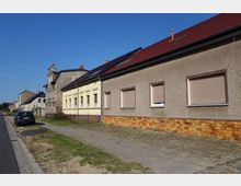 Straßenszene mit einer Reihe niedriger Wohnhäuser, darunter ein beige verputztes Gebäude mit geschlossenen Rollläden und ein gelbes Haus mit verzierten Fenstereinfassungen. Der Gehweg ist aus Pflastersteinen, daneben verläuft eine asphaltierte Straße, auf der links ein Auto geparkt ist.