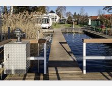 Steg aus Holz, der über ein kleines Gewässer führt, umgeben von Schilf und angelegten Gartenflächen mit einem Wohnhaus und einem Wohnmobil im Hintergrund. Links ein Metallkasten mit einer Laterne auf der Oberseite als Teil der Konstruktion.