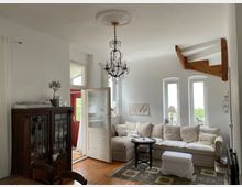 Helles Wohnzimmer mit weißen Wänden und Holzboden. Im Raum befinden sich ein großes cremefarbenes Ecksofa, ein schwarzer Beistelltisch, ein antiker Holzschrank mit Glasfronten, und ein Kronleuchter an der Decke; die Fenster lassen Tageslicht herein.