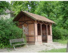 Kleiner Unterstand aus roten Ziegelsteinen mit Holzbalken und Satteldach, umgeben von dichter Vegetation; davor steht eine schlichte Parkbank auf sandigem Boden.