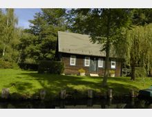 Holzhaus mit Giebeldach in ländlicher Umgebung, umgeben von grüner Wiese, Bäumen und Büschen; im Vordergrund verläuft ein schmaler Wasserkanal mit Holzbefestigung.