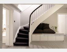 Moderne Inneneinrichtung mit einer weißen Wendeltreppe und einem schwarzen Handlauf. Unter der Treppe befindet sich eine Sitzbank mit schwarzen Kissen und Schubladen, daneben steht eine Vase mit weißen Blumen auf einem grauen Holzboden.